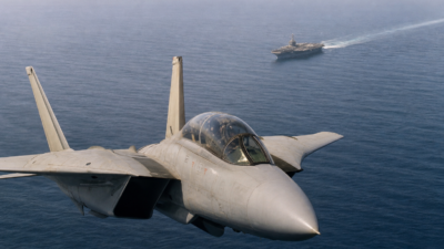 F-14 Tomcat em voo sobre o mar com porta-aviões ao fundo