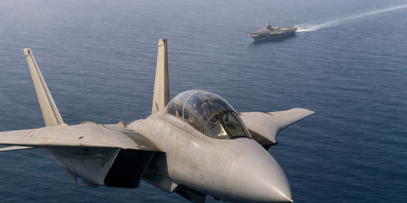 F-14 Tomcat em voo sobre o mar com porta-aviões ao fundo